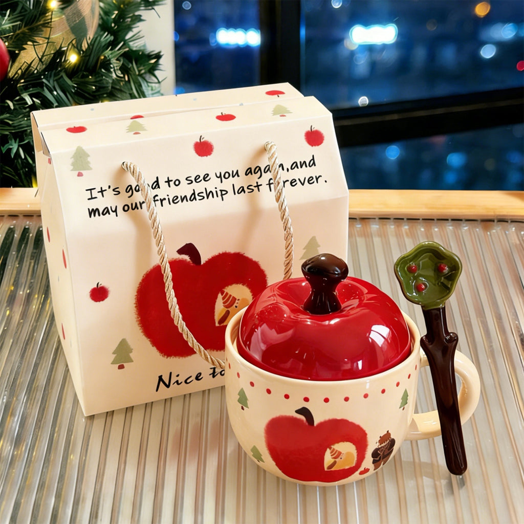 Hand-painted apple-shaped ceramic mug with red apple lid and apple-leaf spoon beside a kraft gift bag printed with red apples and Christmas trees, Christmas tree and evening lights in the background