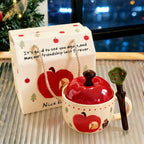 Hand-painted apple-shaped ceramic mug with red apple lid and apple-leaf spoon beside a kraft gift bag printed with red apples and Christmas trees, Christmas tree and evening lights in the background
