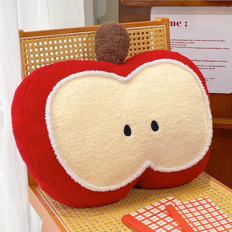 Red apple slice shaped plush pillow with cream white fleece flesh, embroidered seed eyes, and fuzzy brown stem, leaning upright on a rattan chair with an open magazine below