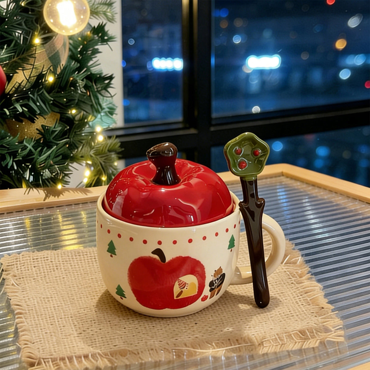 Hand-painted apple-shaped ceramic mug with red apple lid and green apple-leaf spoon, sitting on a burlap coaster near a Christmas tree with warm string lights