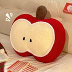 Red apple shaped plush pillow leaning against a cream tufted headboard on a bed, with an open book beside it - cozy reading nook scene showing the plush apple slice design