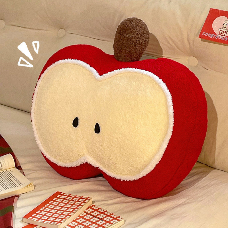 Red apple shaped plush pillow leaning against a cream tufted headboard on a bed, with an open book beside it - cozy reading nook scene showing the plush apple slice design