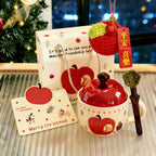 Complete apple mug gift set - hand-painted ceramic mug with red lid, kraft gift bag, knitted apple ornament with a wooden Peace and Joy tag, and a Merry Christmas card, arranged on a table by a Christmas tree