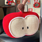 Hand lifting the red apple plush pillow by its fuzzy brown stem, showing the apple-slice face with embroidered seed eyes and soft arctic velvet texture