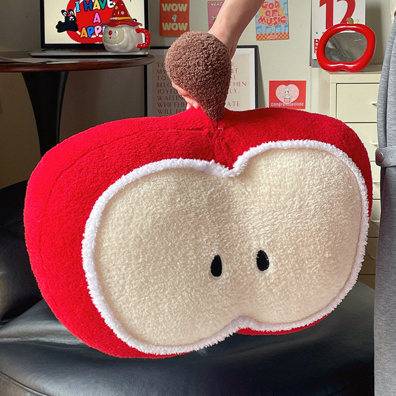 Hand lifting the red apple plush pillow by its fuzzy brown stem, showing the apple-slice face with embroidered seed eyes and soft arctic velvet texture