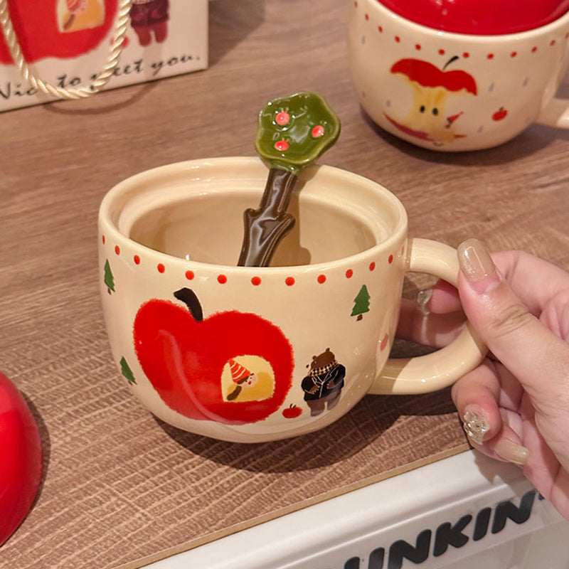 Hand-painted apple ceramic mug held by a woman's hand, showing the red apple illustration and Christmas tree pattern on the body, with the green apple-leaf spoon resting inside