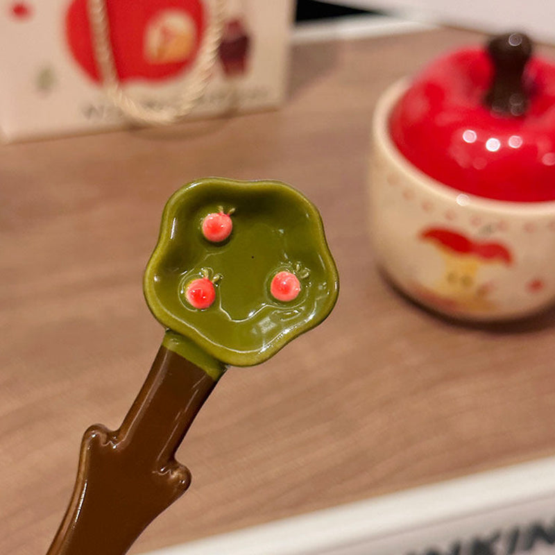 Close-up of the green apple-leaf shaped ceramic spoon with three tiny painted apples on the leaf, held above the apple ceramic mug with red lid