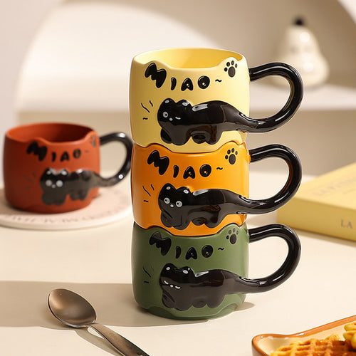 Three black cat ceramic mugs (warm yellow, orange, and dark green) stacked on a wooden desk, each with a 3D relief black cat draped around the body, cat-ear rim, and glossy black tail handle