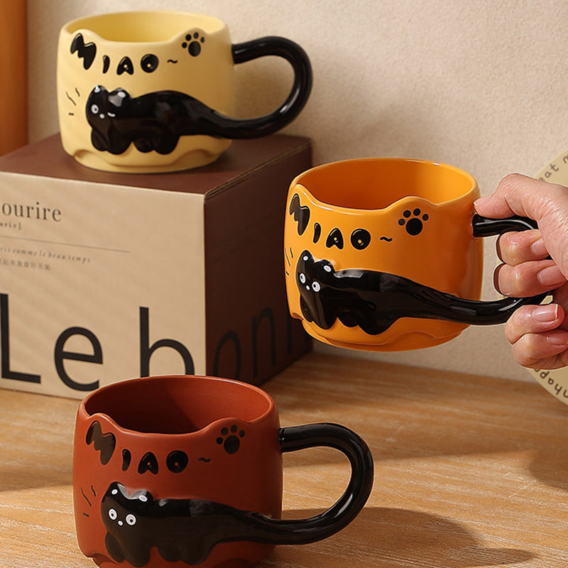 Three black cat ceramic mugs arranged with a kraft gift box - one warm yellow on top of the box, one orange held in hand, and one brick red in front, showing the hand-painted MIAO lettering and paw print