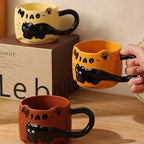 Three black cat ceramic mugs arranged with a kraft gift box - one warm yellow on top of the box, one orange held in hand, and one brick red in front, showing the hand-painted MIAO lettering and paw print