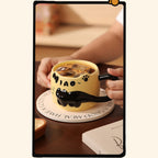 Warm yellow black cat ceramic mug filled with iced coffee, held by two hands on a round wooden coaster on a cafe table, showing the cat-ear rim and glossy tail handle