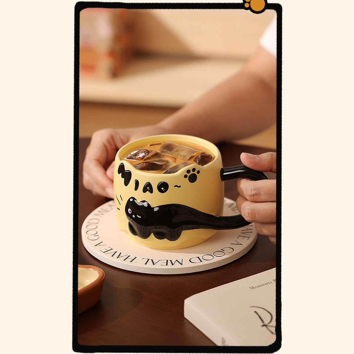 Warm yellow black cat ceramic mug filled with iced coffee, held by two hands on a round wooden coaster on a cafe table, showing the cat-ear rim and glossy tail handle