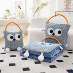 Two blue-check Eye Guy pillow blankets side by side on a living room rug - one folded as a plush monster pillow with giant googly eyes, plush arms and carry handle, the other unzipped and folded out into a full blue flannel blanket, with a white sofa behind