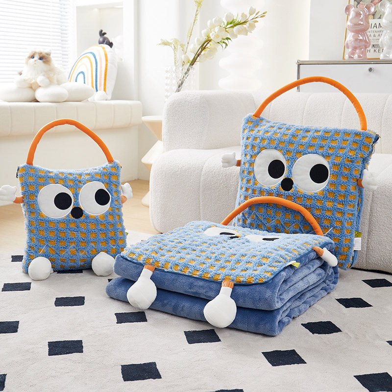 Two blue-check Eye Guy pillow blankets side by side on a living room rug - one folded as a plush monster pillow with giant googly eyes, plush arms and carry handle, the other unzipped and folded out into a full blue flannel blanket, with a white sofa behind