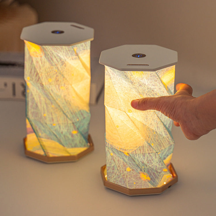 Two lit Starry Night folding lamps on a table - octagonal paper lanterns unfolded into glowing towers printed with Van Gogh's swirling blue, cream and gold sky, white ABS top caps with touch buttons, a hand adjusting the brightness of the nearest lamp