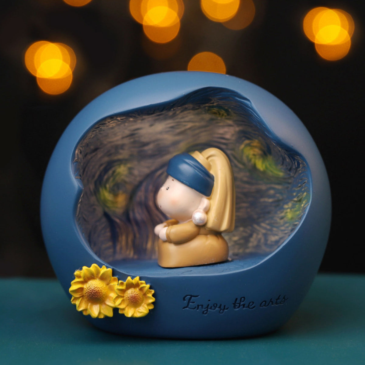 Pearl Earring Girl art night light glowing against warm bokeh lights - matte blue resin sphere carved open to show a Starry Night relief interior, chibi girl with blue headscarf and pearl sitting inside with two sculpted sunflowers and Enjoy the arts lettering