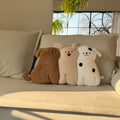 Three long-body dog hug pillows lined up on a cream sofa in golden sunlight - chocolate brown retriever, cream puppy with red tongue out, and spotty white puppy, all with embroidered faces and dangling stitched legs, fern plant and arched window behind