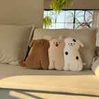 Three long-body dog hug pillows lined up on a cream sofa in golden sunlight - chocolate brown retriever, cream puppy with red tongue out, and spotty white puppy, all with embroidered faces and dangling stitched legs, fern plant and arched window behind