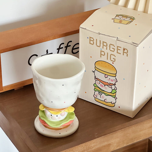 Burger Pig goblet mug beside its dotted gift box on a wooden desk shelf - speckled white ceramic goblet standing on a 3D sculpted burger stem with a rosy-cheeked piggy tucked between lettuce and cheese layers, cream Burger Pig box illustrated with the full burger-pig stack