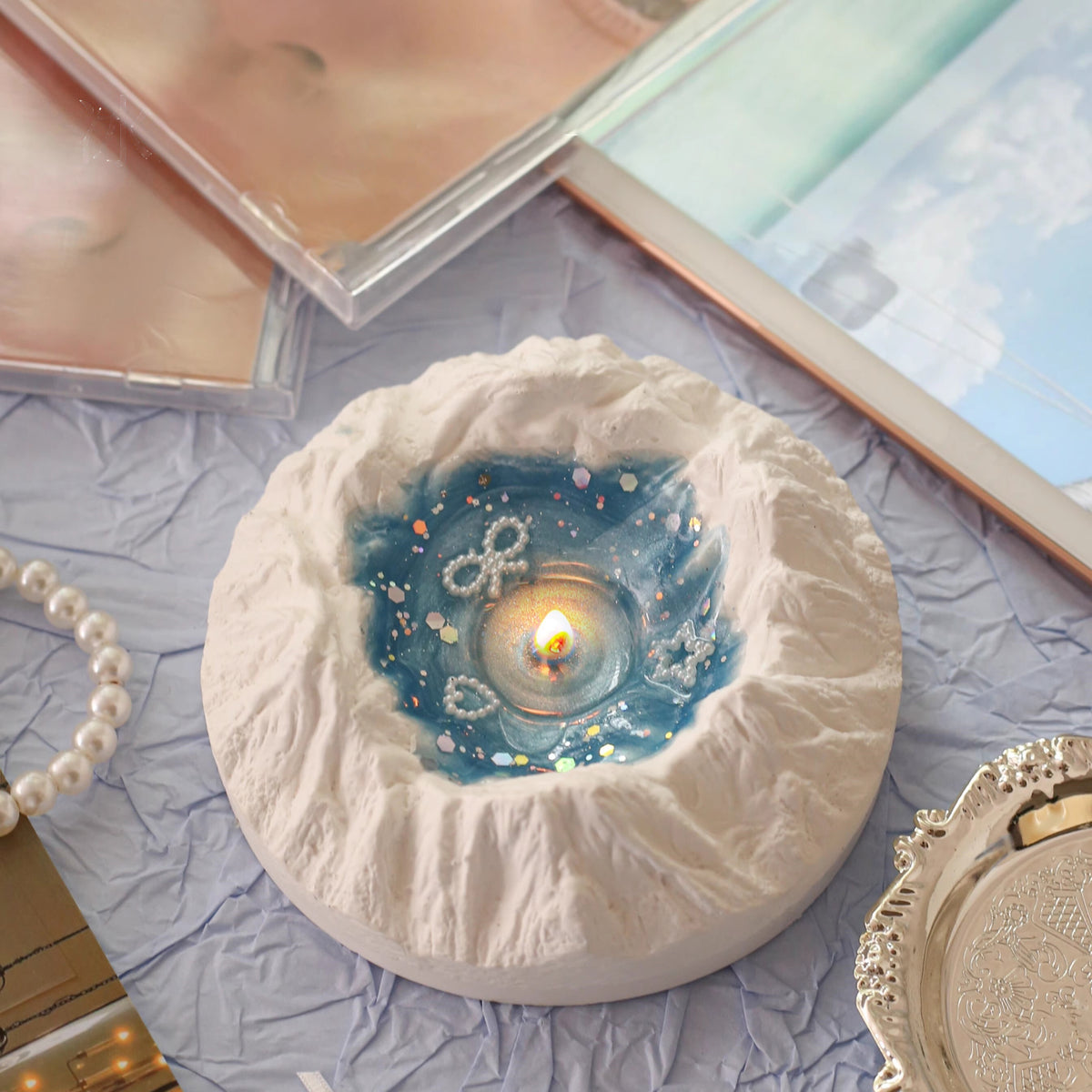 Glacier Ocean candle gift box open - a snow-capped plaster iceberg holding a lit pool of ocean-blue jelly wax with pearl star charms and holographic glitter, nested in light blue tissue paper inside a dusty blue Glacier Candles gift box