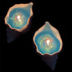 Two lit Fireworks at Sea shell candles against a black background - sculpted pearlescent conch shells filled with ocean-blue jelly wax, each with a holographic firework of colorful sparkles glowing around the flame