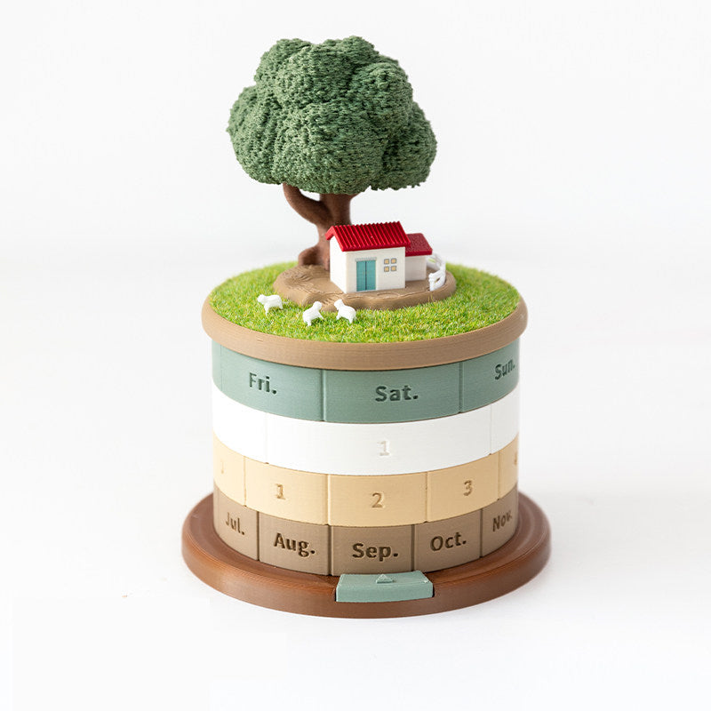 Treehouse perpetual calendar on a white background - 3D printed rotating desk calendar with a fluffy green tree, red-roofed cottage and three tiny sheep on a grass meadow lid, four hand-turned rings showing Fri Sat Sun, date and months below