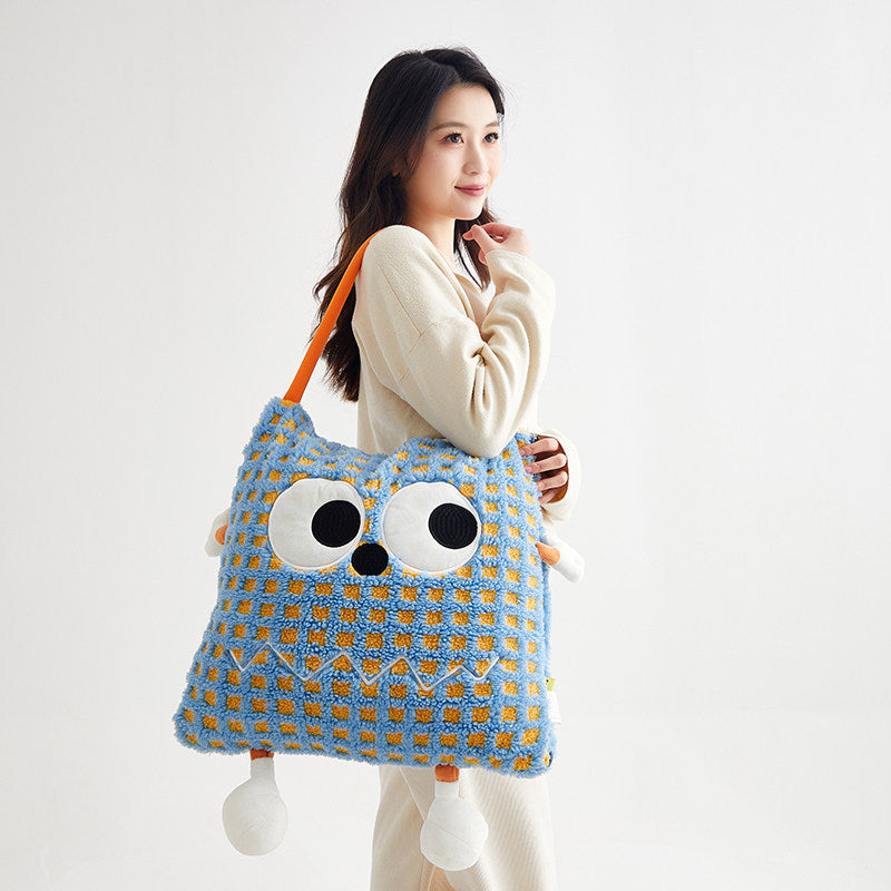 Woman carrying the blue check Eye Guy pillow blanket over her shoulder by its orange handle, showing the monster face with googly eyes, zigzag stitched mouth, plush arms and dangly feet - the folded pillow doubles as a quirky handbag
