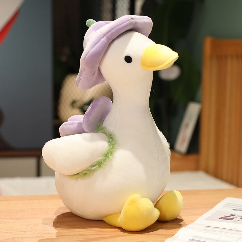 Violet goose plush with lavender-petal flower hat and fuzzy green leaf scarf, sitting on a wooden desk - cream body, embroidered eyes, yellow bill and feet, 40cm kawaii stuffed animal