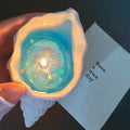 Hand holding a lit Fireworks at Sea shell candle - a pearlescent conch shell with glowing ocean-blue jelly wax and holographic firework sparkles inside, next to a white card that reads Have a nice day