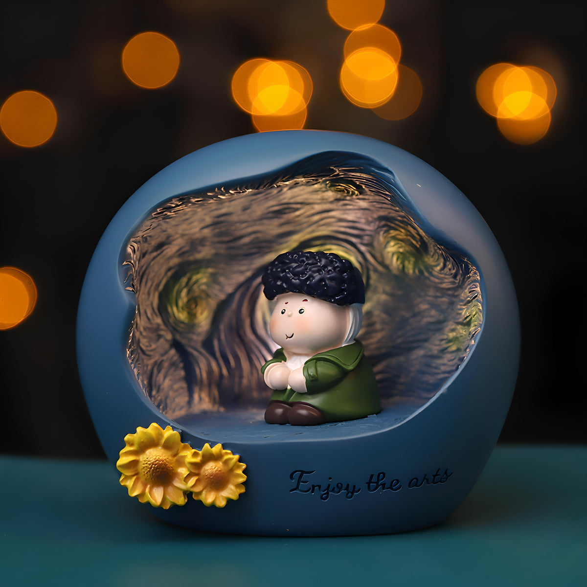 Van Gogh art night light glowing against warm bokeh lights - matte blue resin sphere with Starry Night swirl relief inside, chibi Van Gogh with dark curly hair and green coat sitting cross-legged, two yellow sunflowers and Enjoy the arts lettering on the base