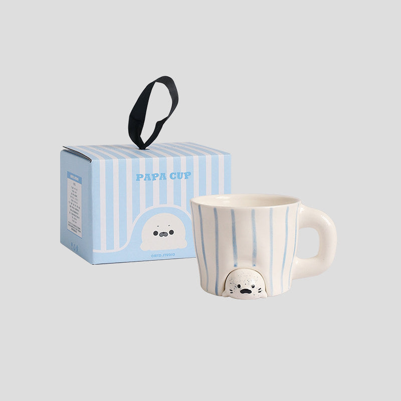 Peekaboo seal mug with its blue-striped gift box on a light grey background - hand-painted white ceramic mug with soft blue stripes, a speckled 3D baby seal peeking out from under the base, chunky rounded handle, striped gift box with ribbon handle