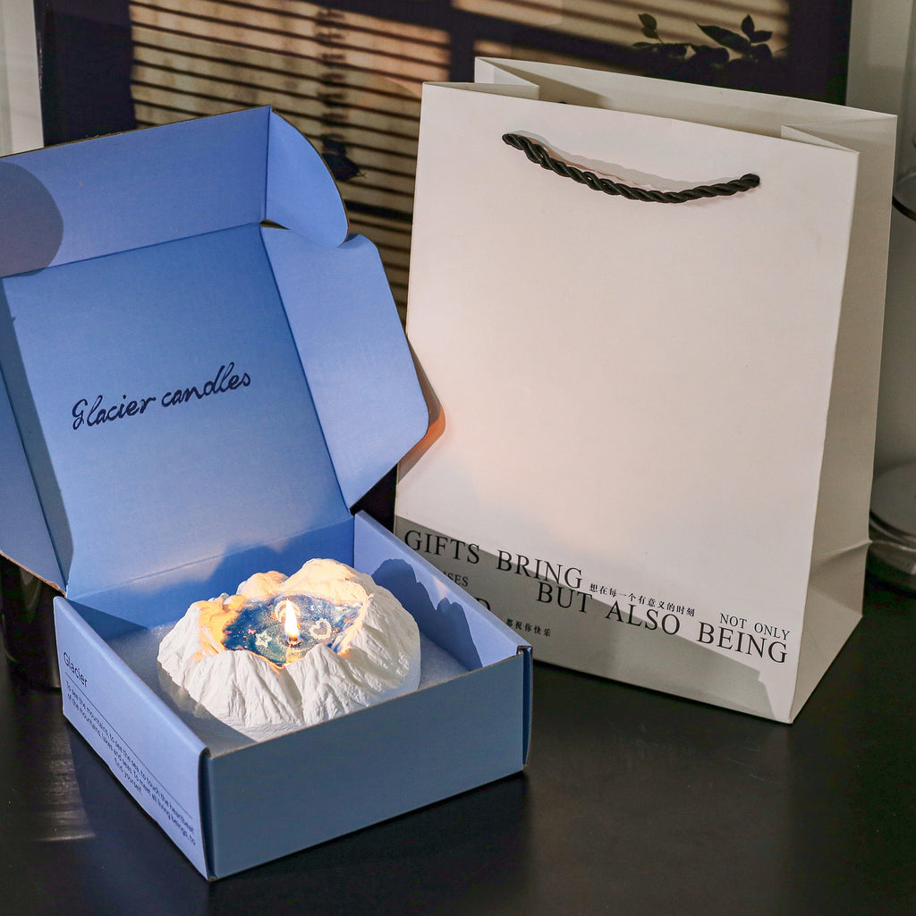 Glacier Ocean candle gift set - dusty blue Glacier Candles gift box open showing the lit plaster iceberg candle with glowing blue jelly wax, beside a white gift bag with rope handle printed with Gifts Bring But Also Being