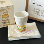 Burger Pig goblet mug resting on a folded cream cloth - speckled white ceramic bowl on the 3D sculpted burger stem with piggy face, lettuce, tomato and bun layers, gift box and framed quote in the background