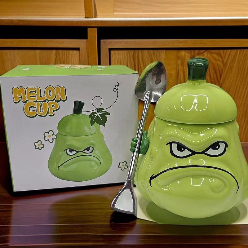 Close-up of the Grumpy Melon Cup gift set - 3D green melon mug with furrowed brows and downturned frown, stem lid on top, shovel spoon hanging on the rim, beside the Melon Cup gift box with cartoon melon illustration