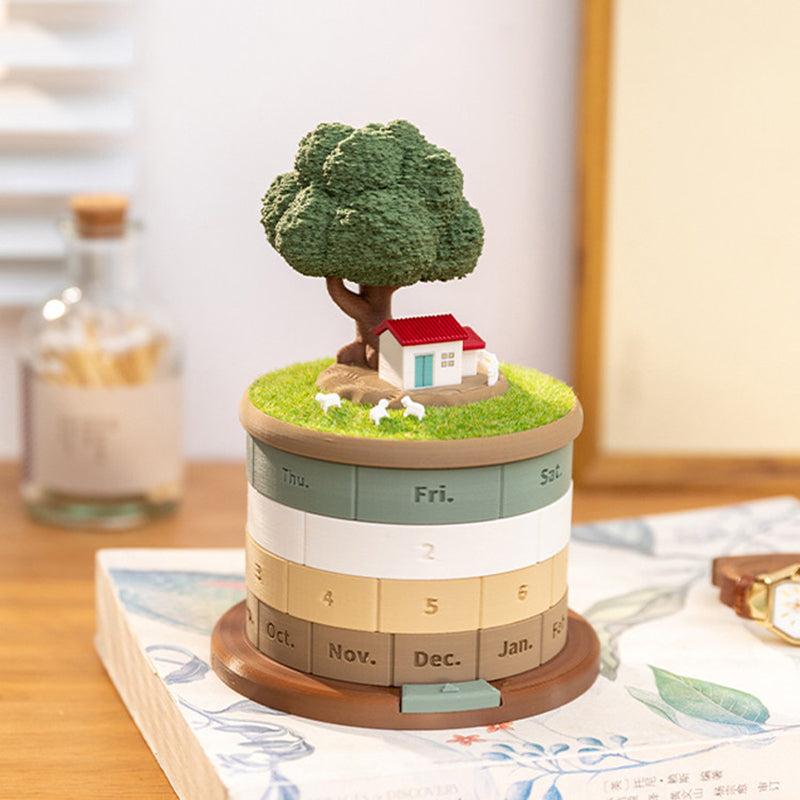 Treehouse perpetual calendar standing on an open book on a wooden desk - miniature cottage and tree on the grass lid, rotating rings showing Thu Fri Sat, dates 4 5 6 and months Oct Nov Dec Jan, mirror and coffee bottle blurred behind