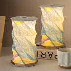 Two Starry Night folding lamps glowing on a white desk beside books and a striped ceramic cup - unfolded paper lanterns with impressionist starry sky print and warm LED light, one leaning slightly at an angle