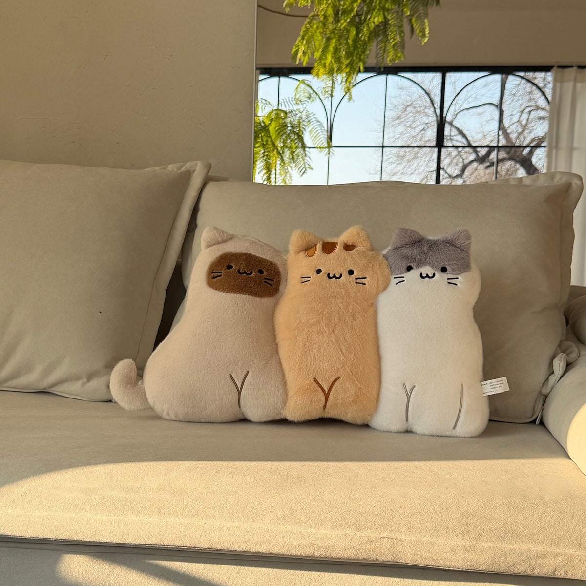 Three long-body cat hug pillows lined up on a cream sofa in golden sunlight - cow-cat, ginger tabby and grey-faced kitty, all with embroidered whisker faces and little stitched tails, fern leaves and window in the background