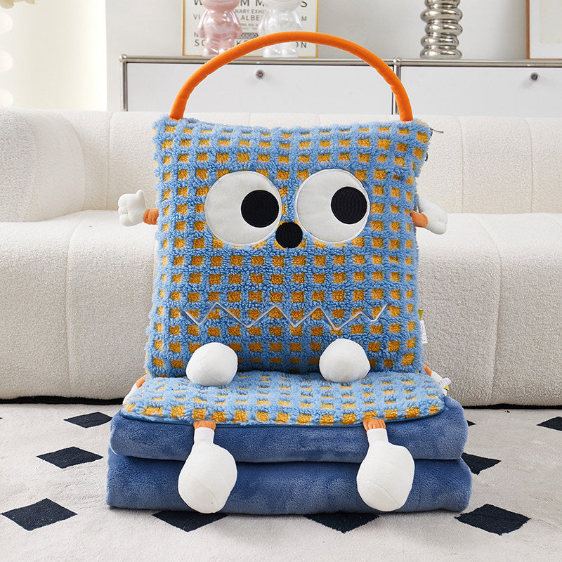 Blue check Eye Guy pillow blanket standing upright on a folded pile of matching blue flannel blanket on a rug, showing the giant googly eyes, wavy stitched grin, plush arms, dangly feet, and orange carry handle