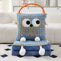 Blue check Eye Guy pillow blanket standing upright on a folded pile of matching blue flannel blanket on a rug, showing the giant googly eyes, wavy stitched grin, plush arms, dangly feet, and orange carry handle
