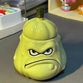Grumpy melon ceramic mug with its stem lid on, standing on a grey desk beside stationery holders - glossy green glaze, 3D sculpted frowning face with bumpy melon texture