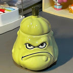 Grumpy melon ceramic mug with its stem lid on, standing on a grey desk beside stationery holders - glossy green glaze, 3D sculpted frowning face with bumpy melon texture