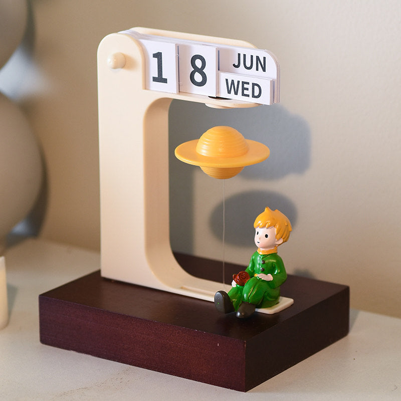 Side view of the floating planet desk calendar - rose prince figurine sitting under the levitating golden Saturn inside the cream flip frame showing 18 JUN WED, on the walnut-grain wooden base