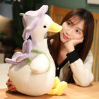 Woman resting her chin on her hand beside the large violet goose plush with lavender flower hat - showing the 40cm size, cream super-soft body and yellow bill on a wooden desk