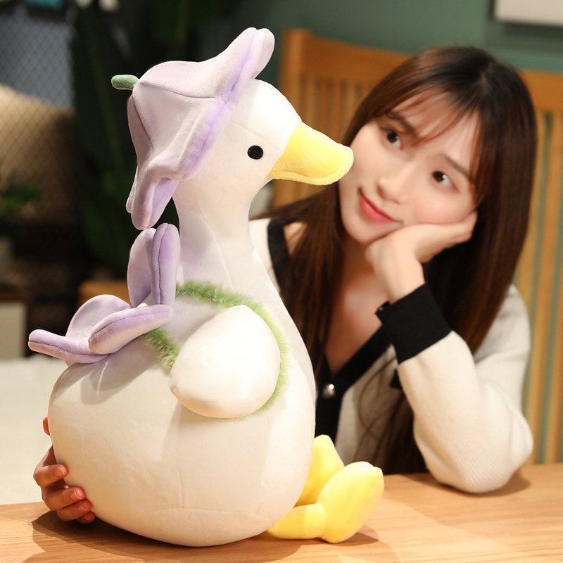 Woman resting her chin on her hand beside the large violet goose plush with lavender flower hat - showing the 40cm size, cream super-soft body and yellow bill on a wooden desk
