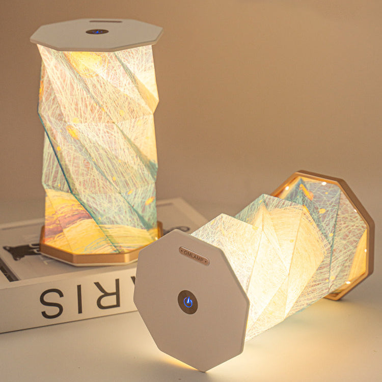 Hand touching the round dimmer button on the octagonal top cap of the Starry Night folding lamp - one lamp fully unfolded and glowing, another mid-fold, showing the accordion paper shade with starry sky print