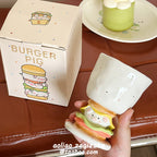 Hand holding the Burger Pig goblet mug - showing the 3D sculpted burger stem with rosy piggy face between lettuce and cheese layers, speckled white ceramic bowl on top, dotted Burger Pig gift box and matcha dessert plate behind