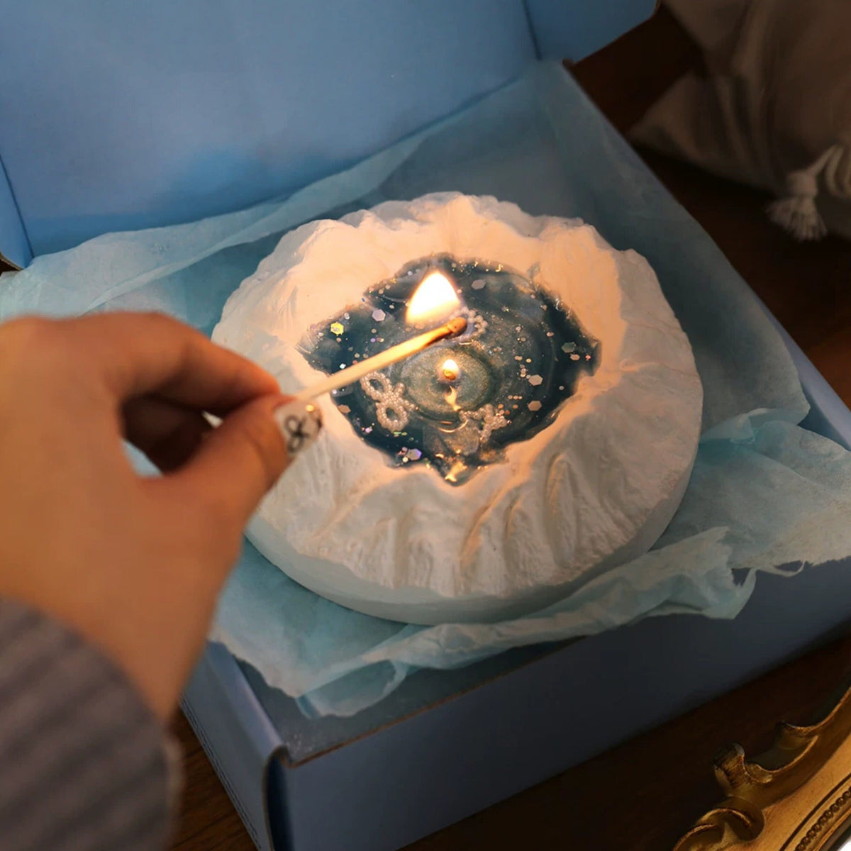 Close-up of the Glacier Ocean candle - snow-white sculpted plaster iceberg cradling a starry ocean-blue jelly wax pool with a lit flame, pearl heart, bow and star charms, holographic hexagon glitter, pearls and gold tray beside