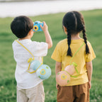 Two children in a green field wearing the gourd water bottles crossbody - boy in white shirt with the blue bottle on a yellow strap, girl in yellow tee with the pink bottle on a green strap, boy holding a toy camera