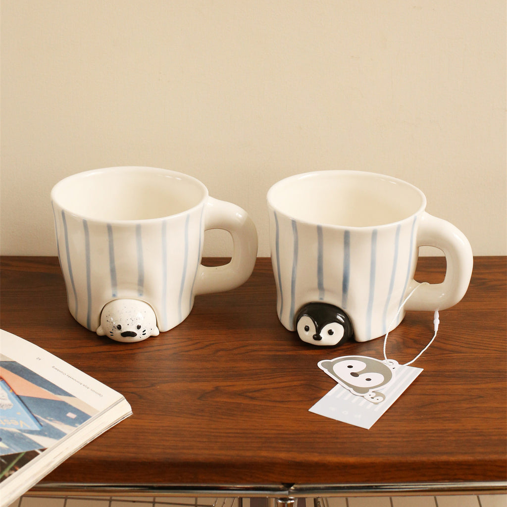 Peekaboo seal and penguin mugs standing side by side on a wooden desk - hand-painted blue striped ceramic cups with the speckled baby seal and glossy penguin peeking out from under each base, an open magazine and illustrated hang tag nearby