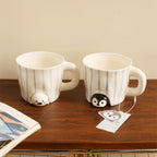 Peekaboo seal and penguin mugs standing side by side on a wooden desk - hand-painted blue striped ceramic cups with the speckled baby seal and glossy penguin peeking out from under each base, an open magazine and illustrated hang tag nearby